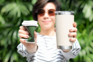 A woman holding a takeaway coffee cup in both hands, one is a single use paper cup with plastic lid the other one is a reusable stainless tumbler - showing you a more sustainable option.