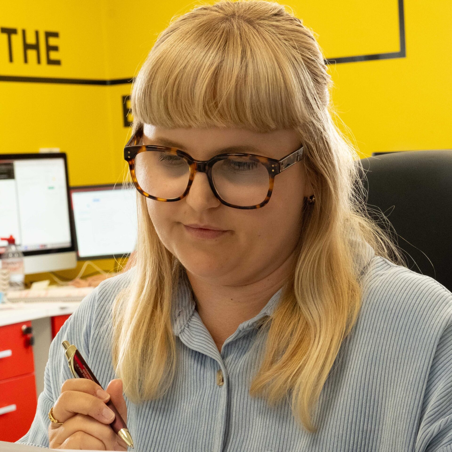 Rosie holding a pen looking to be reading something while sitting In the office with the rest of the team.